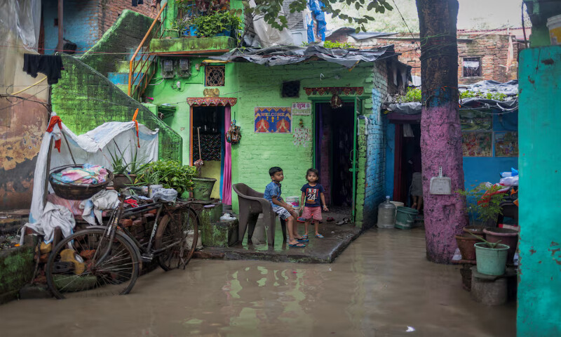 Children sit outside their house next to a flooded alley in New Delhi, India. – Reuters