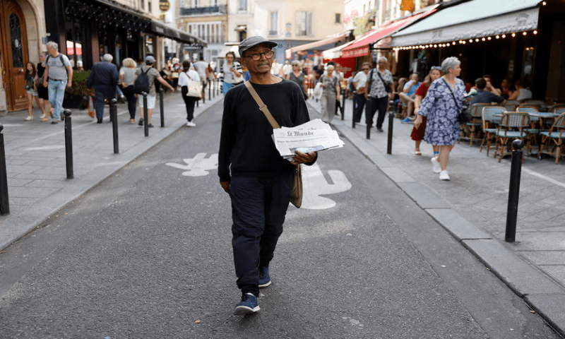 Ali Akbar, 73, known as the last newspaper hawker in the French capital sells ‘Le Monde’ newspaper to people on a terrace in the Latin Quarter, in Paris, on August 4, 2025.
