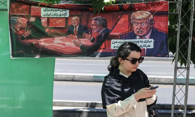 An Iranian woman looks at her mobile phone as she walks past a banner depicting a cartoon of the US president, which reads, 'So we go for the freedom of Iranian women' (left) and 'There is no need for a video, I will do whatever you say' (right), in the capital Tehran on May 9, 2026. AFP